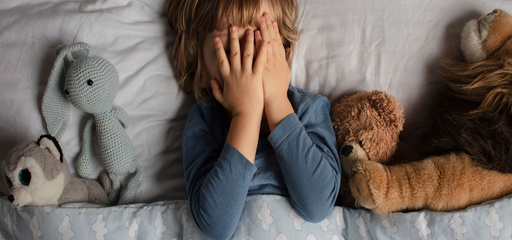 Scared toddler lying in bed with toys and hiding his face 